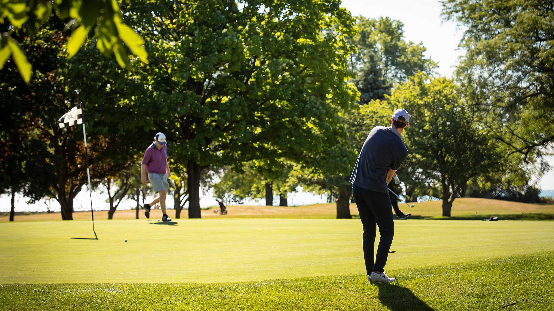 A man chips his golf ball from the fringe onto the green. His friend walks near the pin in the background.