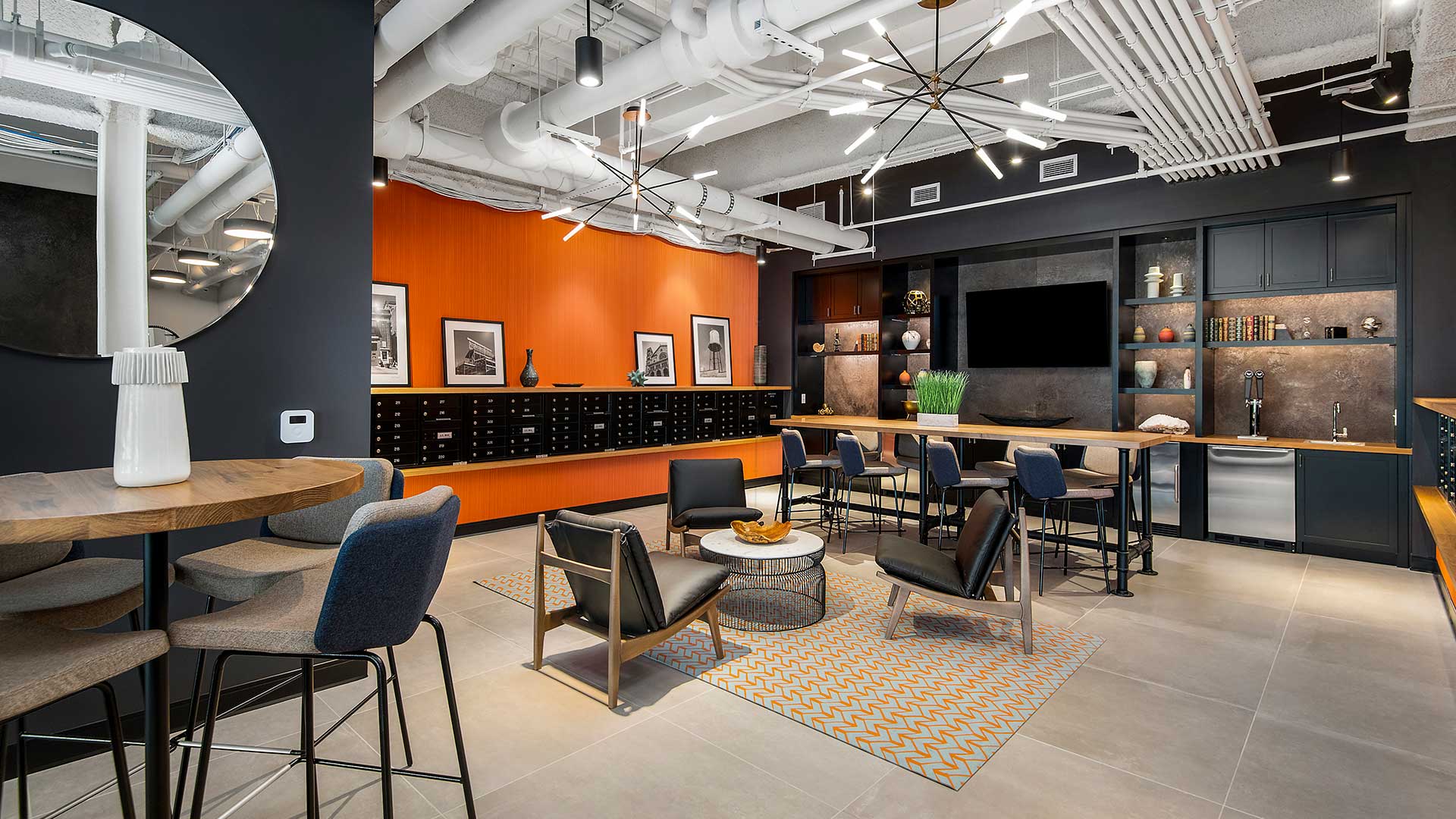 Looking in to the Clubroom at Wrigleyville Lofts. There are club chairs in a circle ahead with a high-top table behind. The far wall has a mounted television with beer taps beside. The side walls are orange with mailboxes built-in for residents.