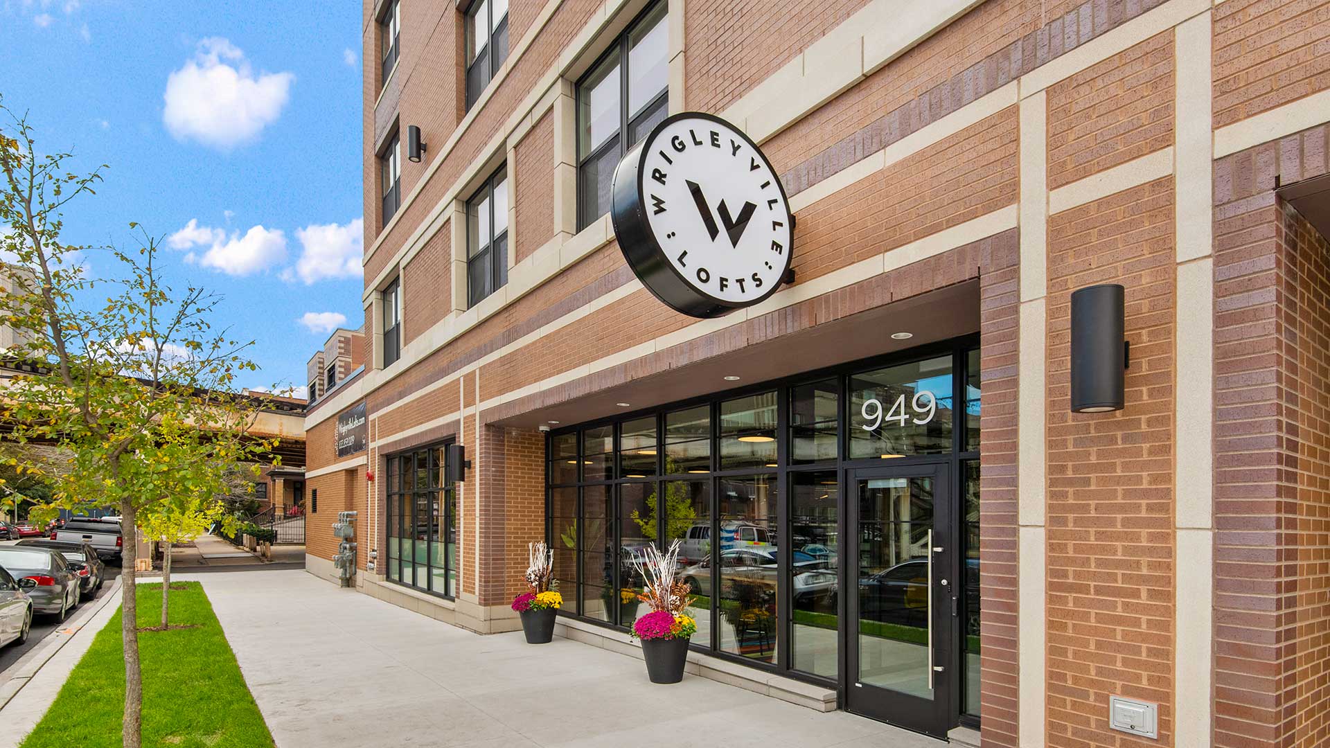 The main entrance to Wrigleyville Lofts along Dakin Street. The elevated train tracks are down the sidewalk to the left, the lobby to the building to the right. The circular Wrigleyville Lofts sign is seen above, mounted to the building.