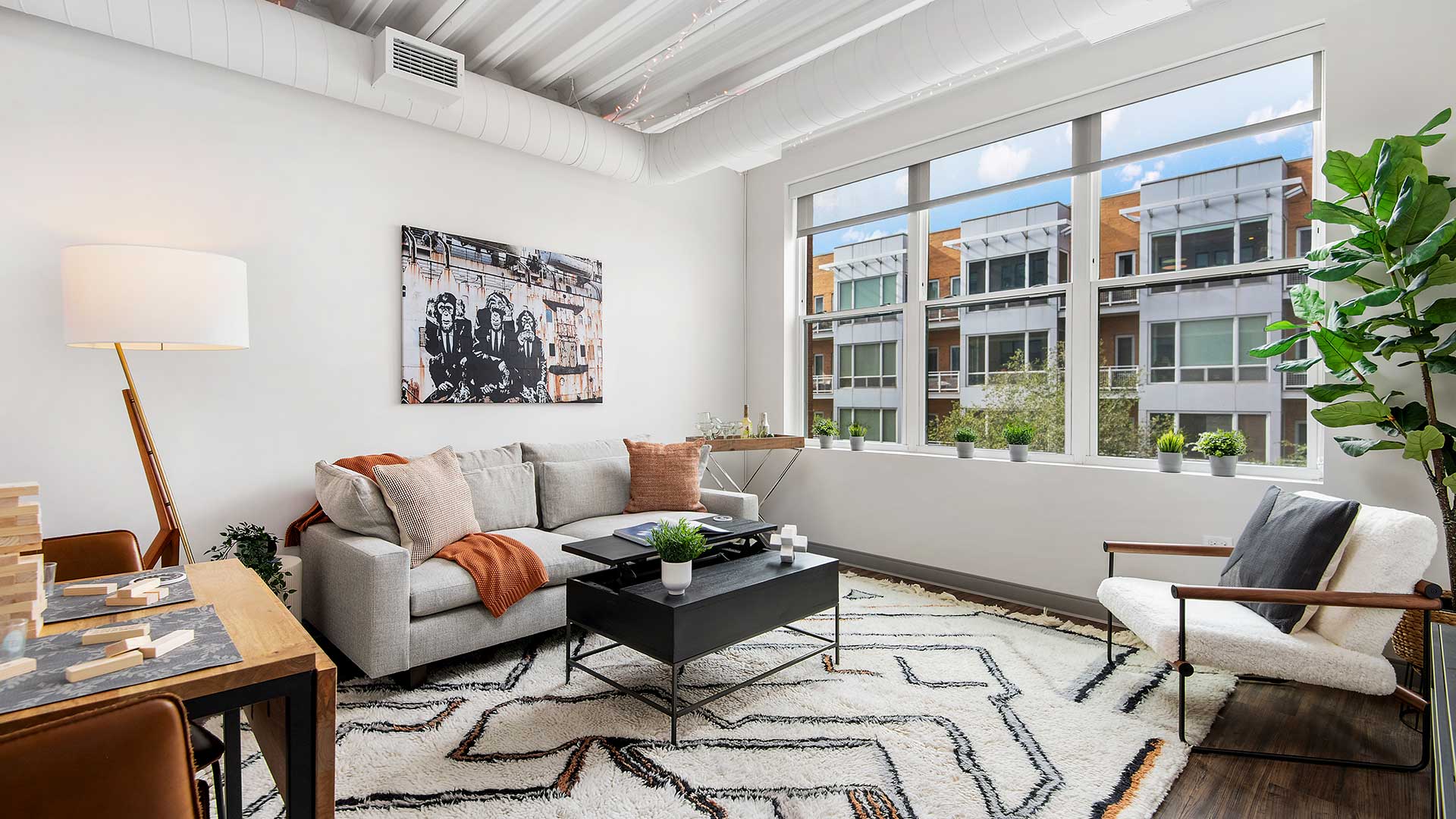 A living room in a residence at Wrigleyville Lofts. A couch and coffee table of off to the left, with a small, sectional table to the near left. Out the windows, to the right, other buildings are seen on a bright day.