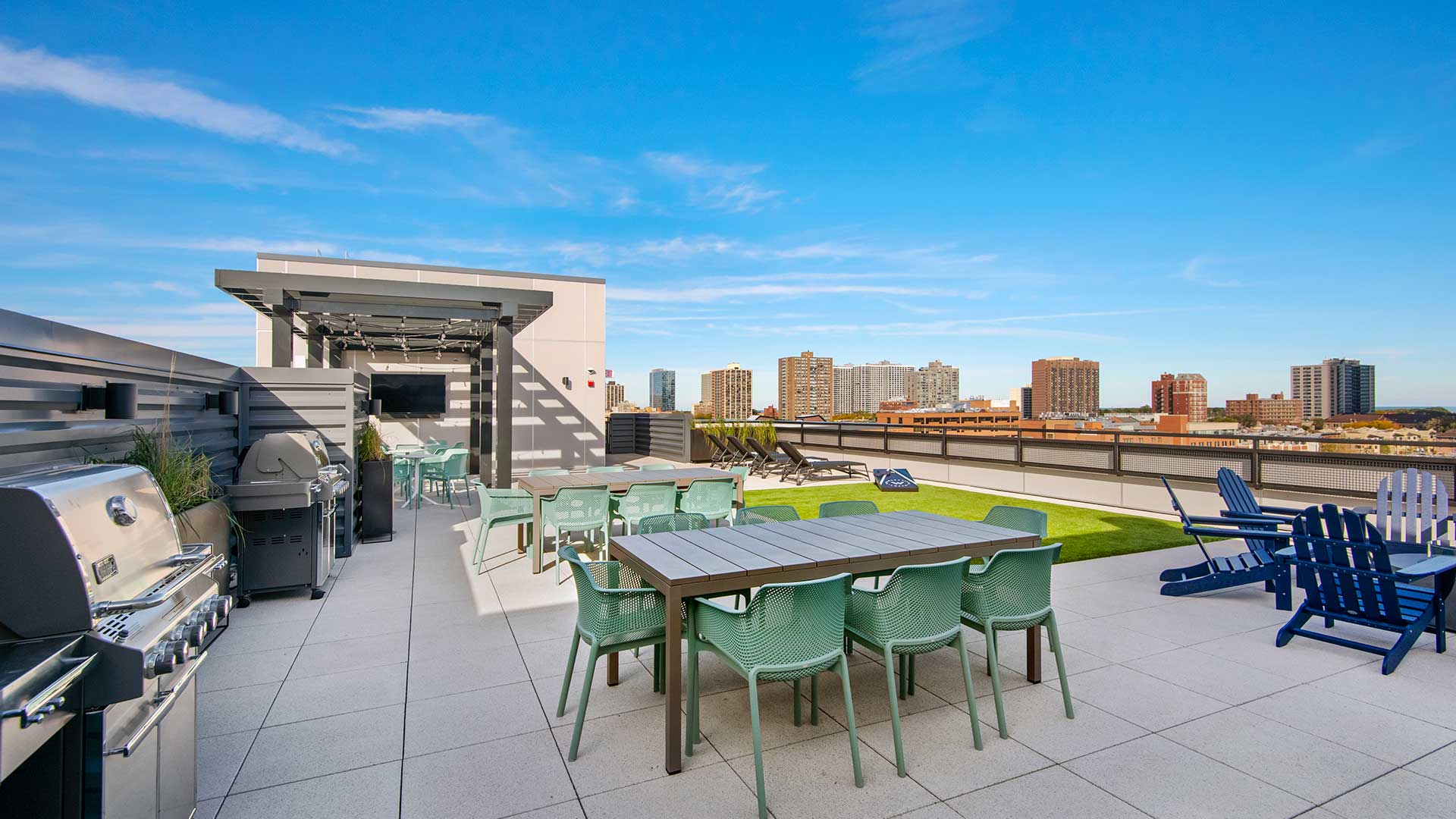 The outdoor kitchen and grilling areas on the rooftop deck at Wrigleyville Lofts. Two outdoor dining tables sit ahead of grills. A television viewing area is behind.