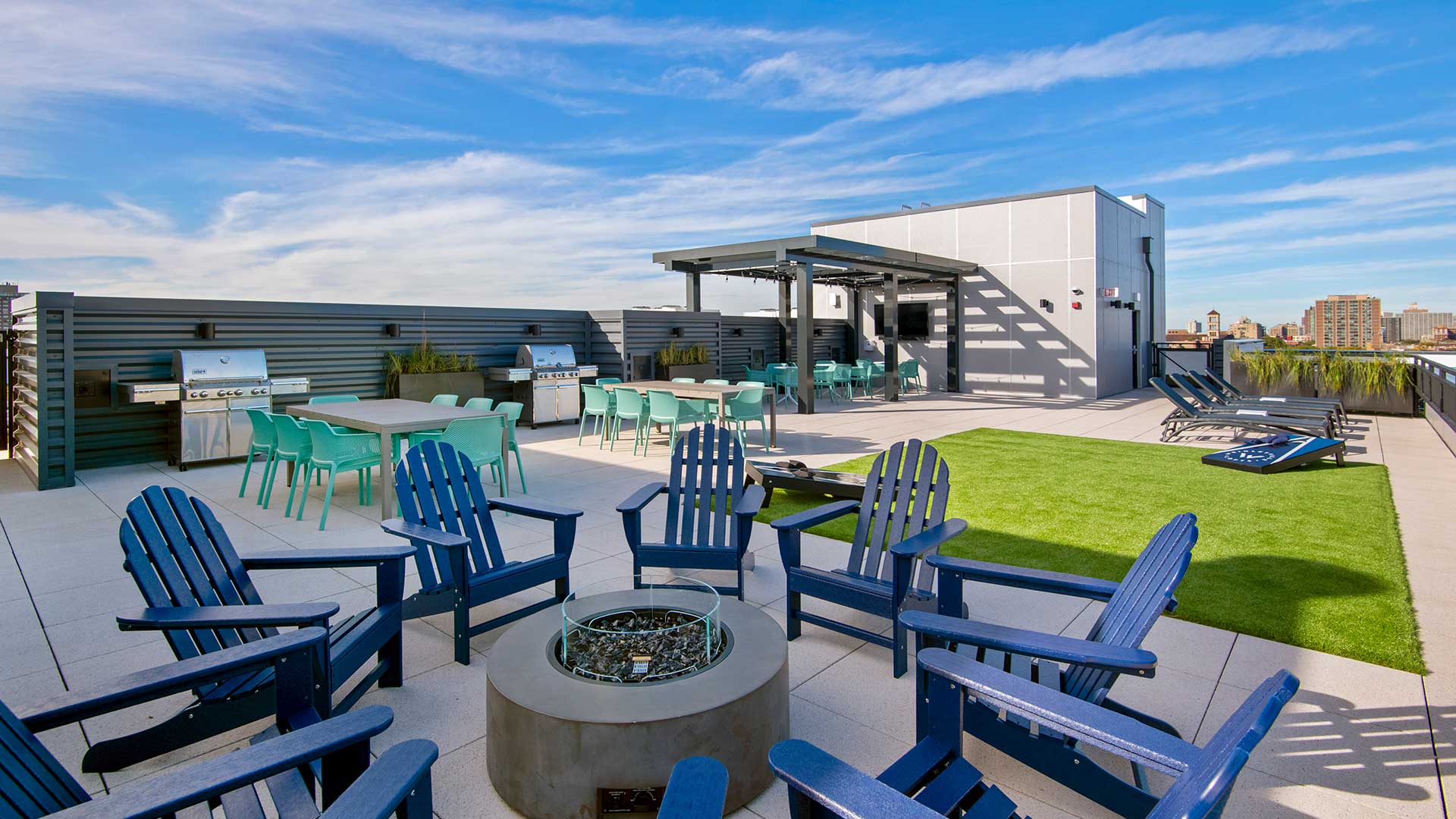 The rooftop deck at Wrigleyville Lofts standing by the fire pit. Chairs circle the fire pit with turf beyond for a bags game. Further back are the grilling areas and television.