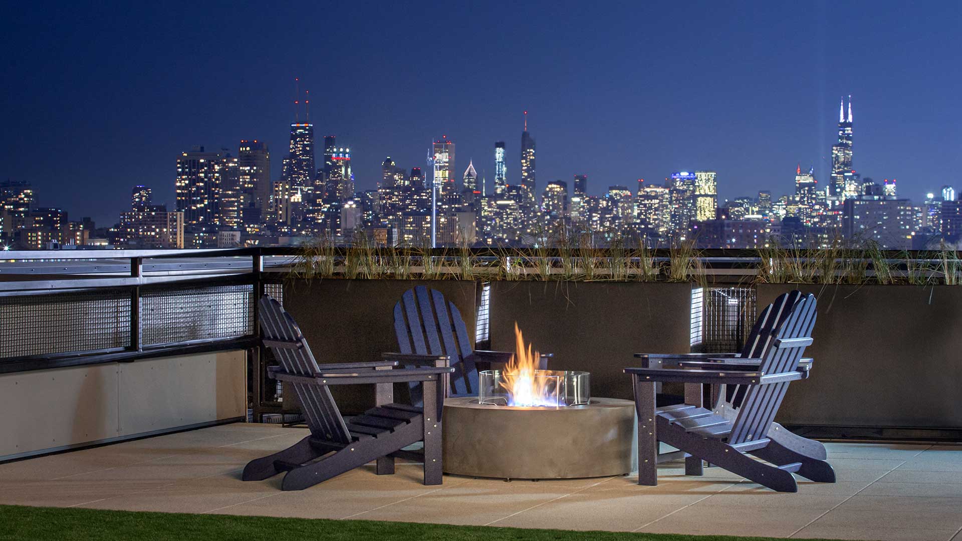 Standing before the fire pit are on the rooftop deck at Wrigleyville Lofts with the Chicago skyline seen in the distance at night.